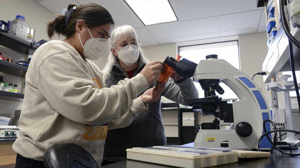 Two women inspect a microscope in a lab.