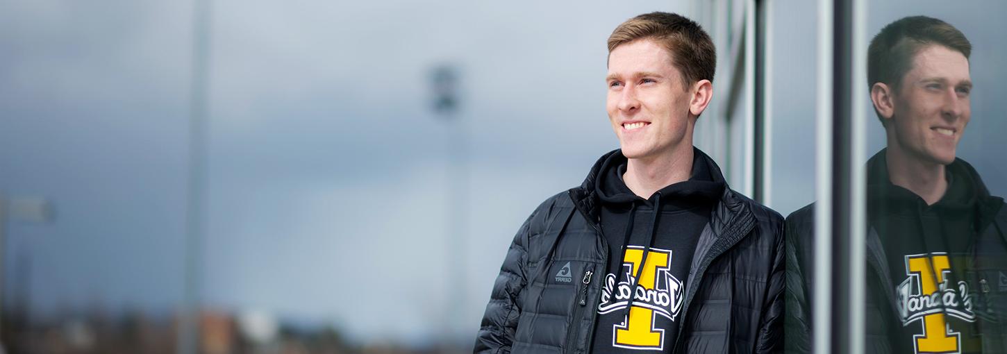 Young man wearing University of Idaho sweatshirt and black jacket leaning against a window at a sport complex.